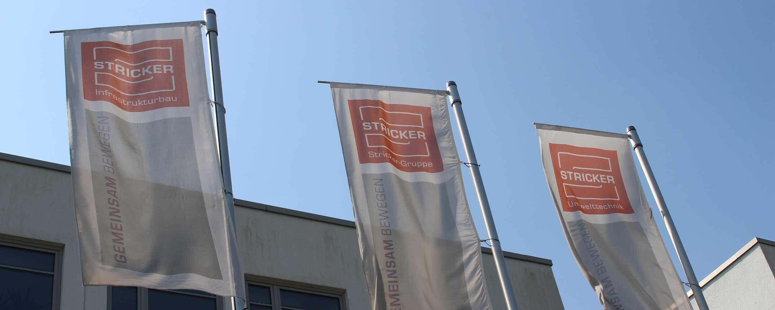 Drei Flaggen mit Firmenlogo vor einem Gebäude der Stricker Gruppe